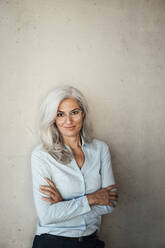 Smiling gray haired businesswoman wearing eyeglasses standing with arms crossed in front of wall - JOSEF08439