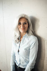 Smiling businesswoman wearing eyeglasses standing in front of wall - JOSEF08440