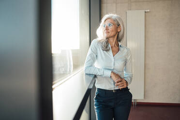 Businesswoman wearing eyeglasses looking through window standing in office - JOSEF08442