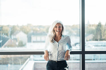 Happy woman with gray hair in front of glass window in office - JOSEF08446