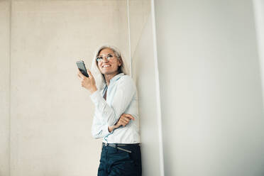 Smiling businesswoman with smart phone standing in front of wall at work place - JOSEF08453