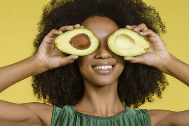 Lächelnde junge Frau, die ihre Augen mit Avocados vor gelbem Hintergrund bedeckt - EIF03584