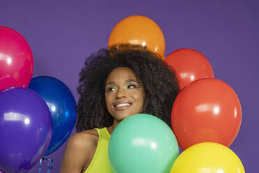 Lächelnde junge Frau mit lockigem Haar inmitten von bunten Luftballons vor lila Hintergrund - EIF03591