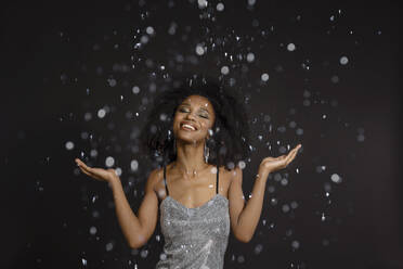 Smiling young woman with eyes closed standing under falling confetti against black background - EIF03605