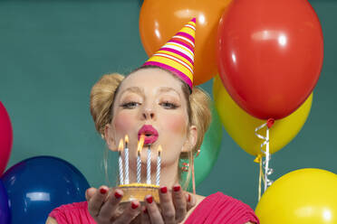 Schöne junge Frau mit Partyhut bläst Kerzen auf Geburtstagskuchen vor grünem Hintergrund aus - EIF03617