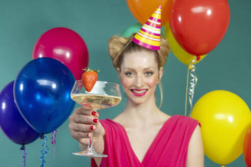 Smiling young woman wearing party hat showing champagne flute with strawberry amidst colorful balloons against green background - EIF03619