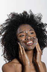 Happy young woman applying scrub facial mask on face against white background - EIF03640