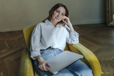 Nachdenkliche Frau mit Laptop auf gelbem Sessel sitzend - OGF01120