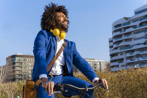 Glücklicher Geschäftsmann mit Kopfhörern und Umhängetasche auf dem Fahrrad - OIPF01460