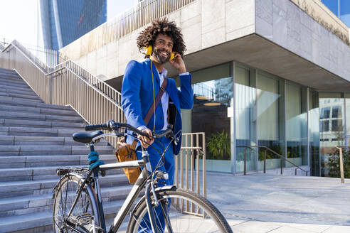 Lächelnder Geschäftsmann mit Fahrrad hört Musik über Kopfhörer an einem sonnigen Tag - OIPF01461