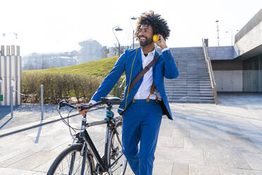 Happy businessman with bicycle listening music through headphones on footpath - OIPF01462