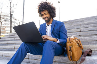 Lächelnder Geschäftsmann, der einen Laptop benutzt und mit einer Umhängetasche auf einer Treppe sitzt - OIPF01463