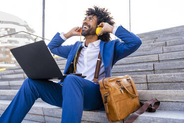 Happy businessman listening music through headphones sitting with laptop on sunny day - OIPF01469