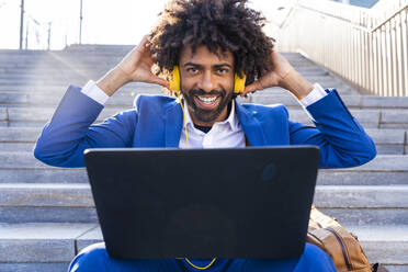 Happy businessman adjusting headphones sitting with laptop on staircase - OIPF01471