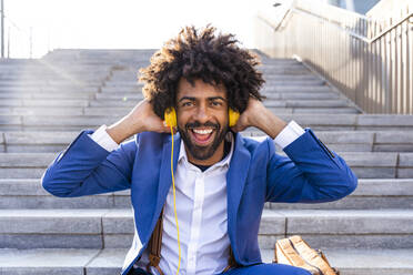 Glücklicher Geschäftsmann, der auf einer Treppe sitzend über Kopfhörer Musik hört - OIPF01478