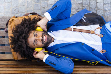 Happy businessman listening music through headphones lying on bench - OIPF01480