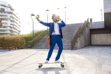 Glücklicher Geschäftsmann mit Einwegbecher, der Musik hört und auf dem Fußweg Skateboard fährt - OIPF01484