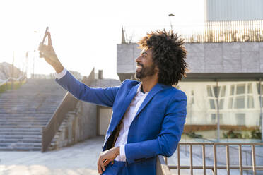 Glücklicher Geschäftsmann, der ein Selfie mit seinem Mobiltelefon macht und sich auf ein Geländer stützt - OIPF01487