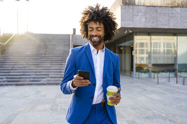 Smiling businessman holding disposable cup using mobile phone walking on footpath - OIPF01492