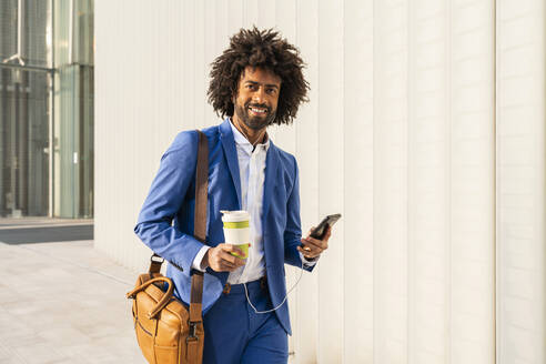 Geschäftsmann mit Smartphone und Einwegbecher geht mit Umhängetasche an der Wand spazieren - OIPF01494