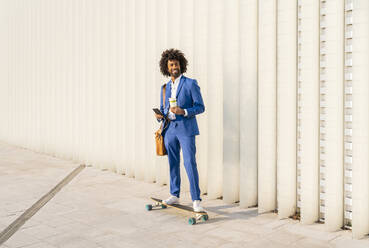 Smiling businessman holding smart phone and disposable cup standing on skateboard in front of wall - OIPF01496