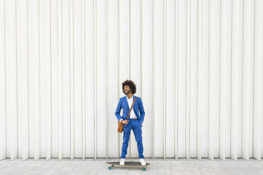 Glücklicher Geschäftsmann mit Umhängetasche auf einem Skateboard vor einer Wand stehend - OIPF01497