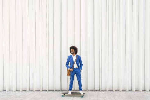 Glücklicher Geschäftsmann mit Umhängetasche auf einem Skateboard vor einer Wand stehend - OIPF01497