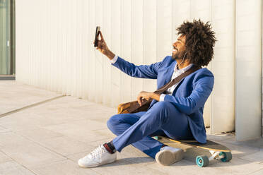 Smiling businessman taking selfie through smart phone sitting on skateboard in front of wall - OIPF01500