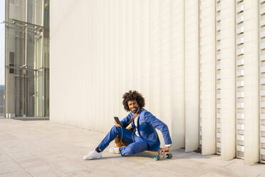 Lächelnder Geschäftsmann mit Mobiltelefon auf einem Skateboard vor einer Wand sitzend - OIPF01501
