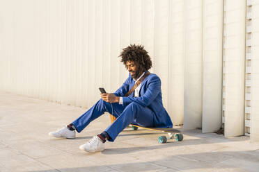 Geschäftsmann, der ein Smartphone benutzt und auf einem Skateboard vor einer Wand sitzt - OIPF01502
