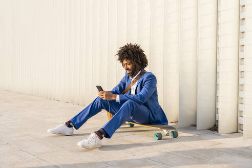 Businessman using smart phone sitting on skateboard in front of wall - OIPF01502