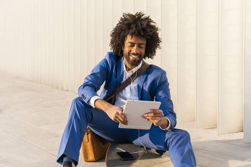 Lächelnder Geschäftsmann mit Mobiltelefon und Tablet-PC auf einem Skateboard sitzend - OIPF01503