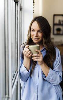 Lächelnde schöne Frau mit Kaffeetasse, die zu Hause am Fenster steht - GIOF15006