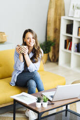 Happy freelancer holding coffee cup sitting on sofa at home - GIOF15018