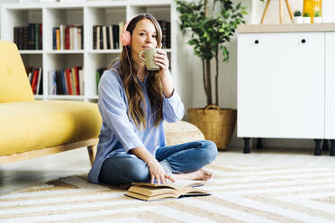Woman having coffee sitting with book on carpet at home - GIOF15022
