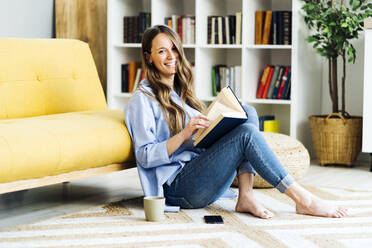 Smiling young woman with book sitting on carpet at home - GIOF15025