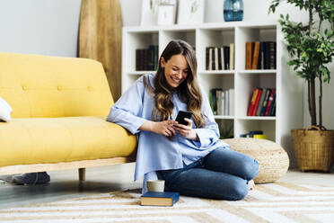 Happy woman using smart phone sitting on carpet by sofa at home - GIOF15026