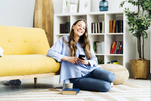 Glückliche Frau mit Smartphone, die zu Hause auf dem Teppich sitzt - GIOF15027