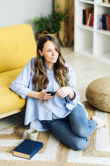 Nachdenkliche Frau mit Smartphone sitzt zu Hause auf dem Teppich - GIOF15028