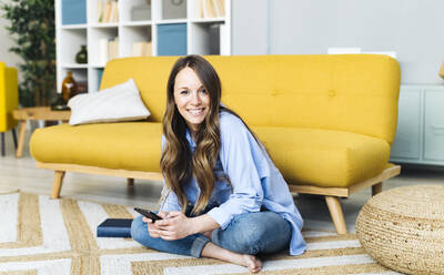 Smiling woman with mobile phone sitting on carpet in living room at home - GIOF15030