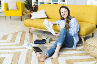 Happy freelancer with laptop and document sitting on carpet by sofa at home - GIOF15033