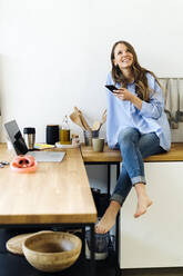 Thoughtful freelancer sitting on kitchen counter with mobile phone at home - GIOF15038