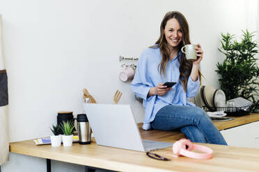 Smiling freelancer sitting on kitchen counter with mobile phone at home - GIOF15039