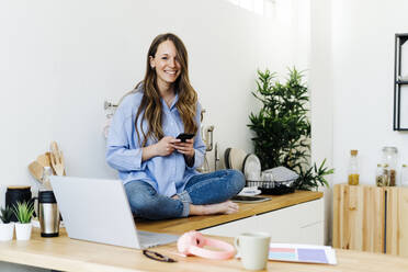 Smiling freelancer with cross-legged sitting on kitchen counter at home - GIOF15041