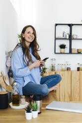 Smiling woman with cross-legged sitting on kitchen counter at home - GIOF15042