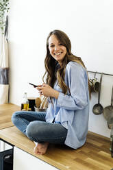 Glückliche Frau sitzt im Schneidersitz auf dem Küchentisch zu Hause - GIOF15043
