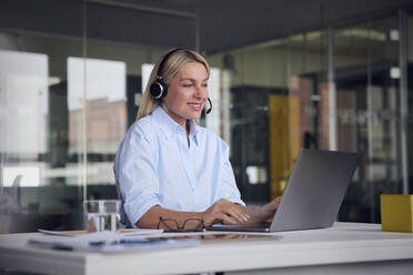 Lächelnde Geschäftsfrau mit Headset und Laptop am Schreibtisch im Büro - RBF08792