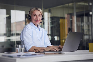 Lächelnde Geschäftsfrau mit Headset und Laptop am Schreibtisch im Büro - RBF08793