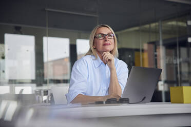 Lächelnde Geschäftsfrau mit Brille und Laptop am Schreibtisch im Büro - RBF08797