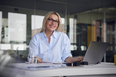 Happy businesswoman holding paper and laptop at desk in office - RBF08800